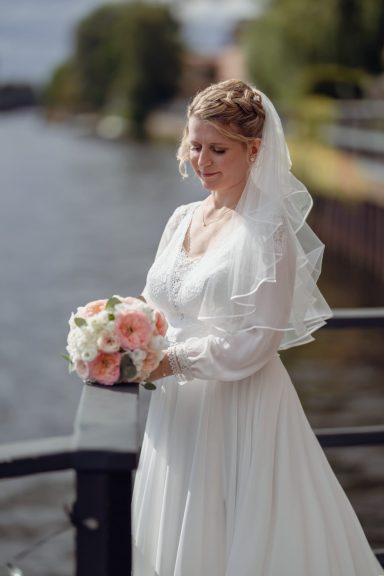 Braut mit Blumenstrauß am Flussufer, trägt ein elegantes weißes Kleid.