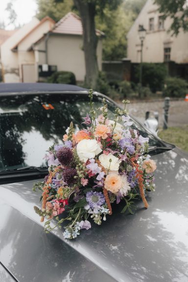 Bunter Blumenstrauß auf dem Dach eines silbernen Autos, umgeben von grüner Landschaft.