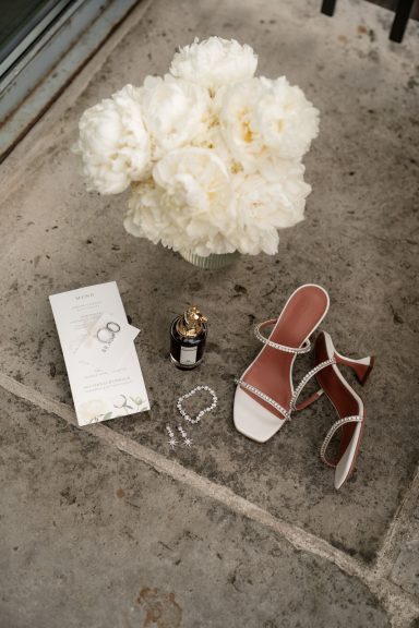 Weiße Blüten, elegante Sandalen, Parfümflasche und Schmuck auf einem grauen Untergrund.