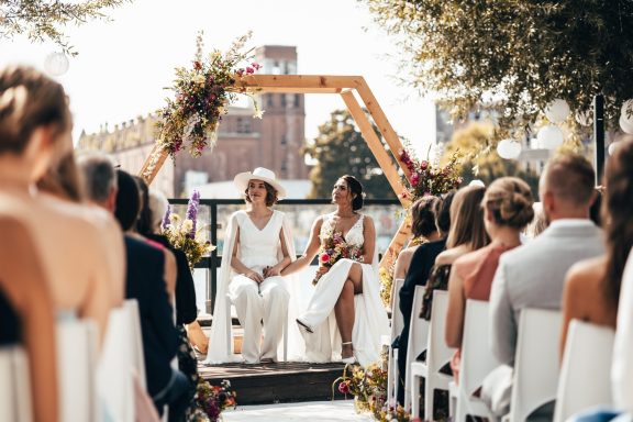 Hochzeit mit Brautpaar unter einem blühenden Bogen, Gäste sitzen im Freien.