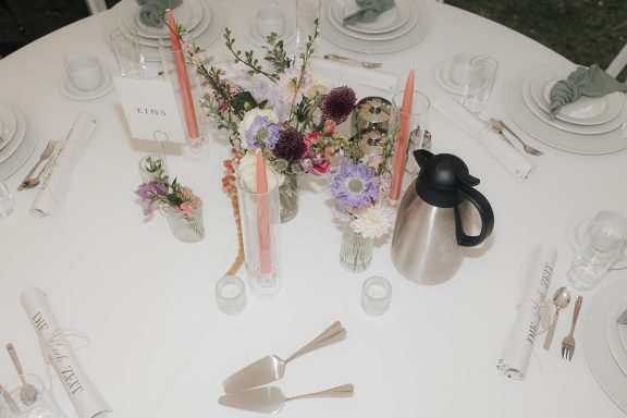 Tisch gedeckt mit Blumenarrangements, Kaffeekanne und Besteck.