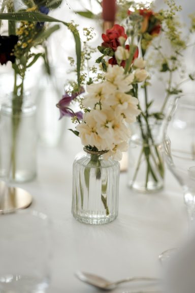 Ein Glas mit weißen Blumen, umgeben von weiteren Blumen in Gläsern, auf einem Tisch.