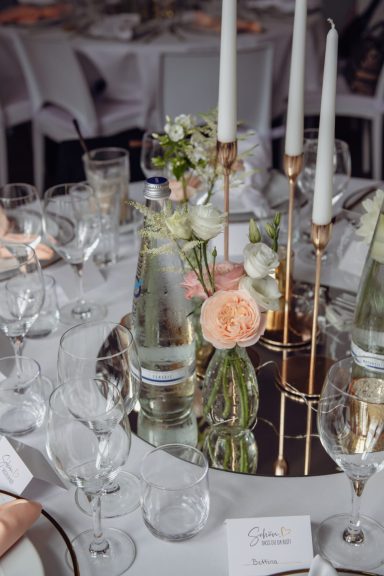 Elegante Tischdekoration mit Kerzenhaltern, Blumen und Gläsern auf einem runden Spiegel.